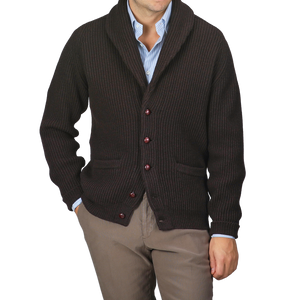 A person dressed in a William Lockie Ebony Brown Lambswool Shawl Collar Cardigan featuring leather buttons, styled over a light blue shirt and teamed with brown pants.