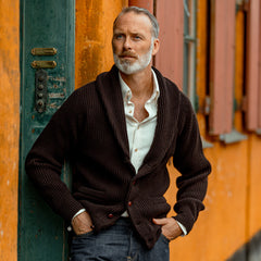 A man leaning against a wall wearing a William Lockie Ebony Brown Lambswool Shawl Collar Cardigan.