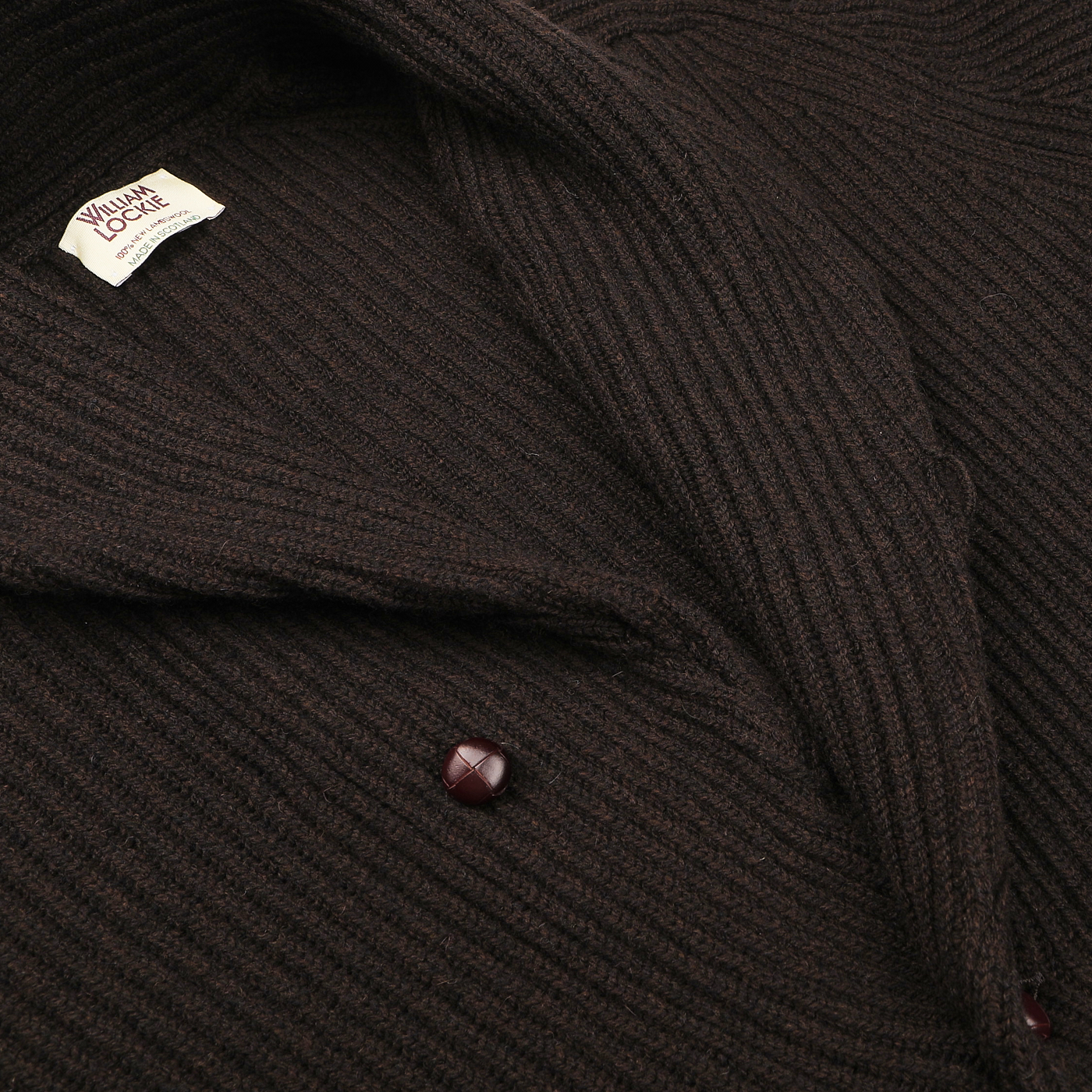 A close-up of the Ebony Brown Lambswool Shawl Collar Cardigan showcases its craftsmanship using Scottish lambswool. The piece, labeled "William Lockie," is elegantly designed with a collar and dark leather buttons.