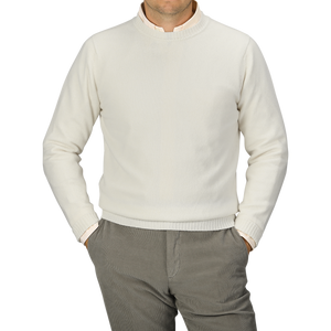 A person is wearing an Ecru Crew Neck Lambswool Sweater from William Lockie and light gray pants, standing with hands in pockets. The person's head is not visible. The background is plain gray, showcasing the elegance of quality knitwear.