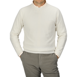 A person is wearing an Ecru Crew Neck Lambswool Sweater from William Lockie and light gray pants, standing with hands in pockets. The person's head is not visible. The background is plain gray, showcasing the elegance of quality knitwear.