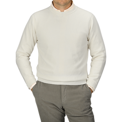 A person is wearing an Ecru Crew Neck Lambswool Sweater from William Lockie and light gray pants, standing with hands in pockets. The person's head is not visible. The background is plain gray, showcasing the elegance of quality knitwear.