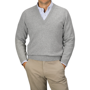 A person wearing a Flannel Grey Deep V-Neck Lambswool Sweater by William Lockie over a collared shirt and beige pants stands against a plain background.