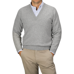 A person wearing a Flannel Grey Deep V-Neck Lambswool Sweater by William Lockie over a collared shirt and beige pants stands against a plain background.