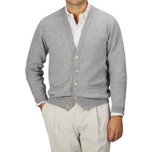 A man is wearing the Flannel Grey Lambswool Saddle Shoulder Cardigan by William Lockie over a white shirt. Paired with light beige pants, his hands are in his pockets as he poses against a plain gray background.