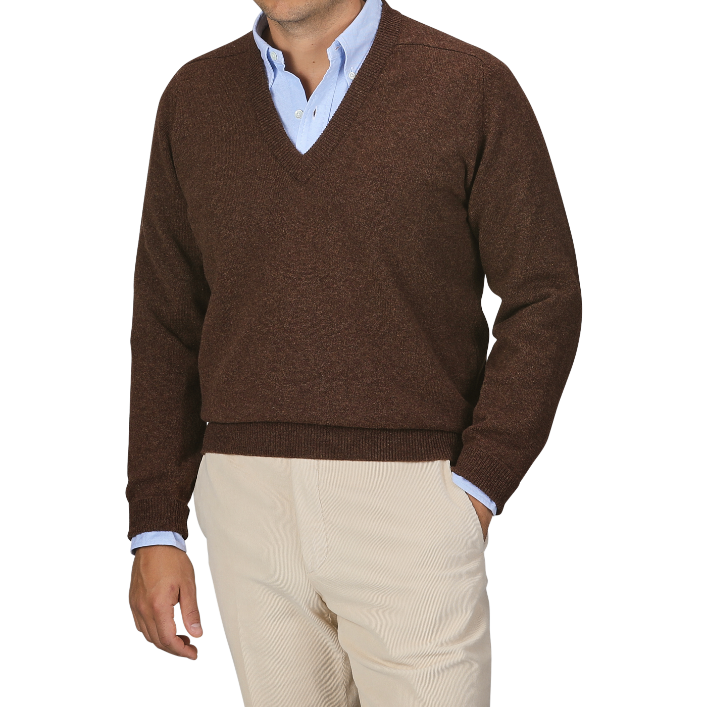 A person dressed in a William Lockie Hickory Brown Deep V-Neck Lambswool Sweater layered over a light blue collared shirt and paired with beige pants stands against a plain background.