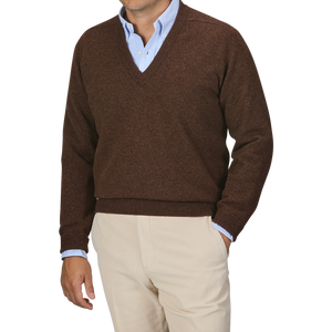 A person dressed in a William Lockie Hickory Brown Deep V-Neck Lambswool Sweater layered over a light blue collared shirt and paired with beige pants stands against a plain background.