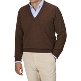 A person dressed in a William Lockie Hickory Brown Deep V-Neck Lambswool Sweater layered over a light blue collared shirt and paired with beige pants stands against a plain background.