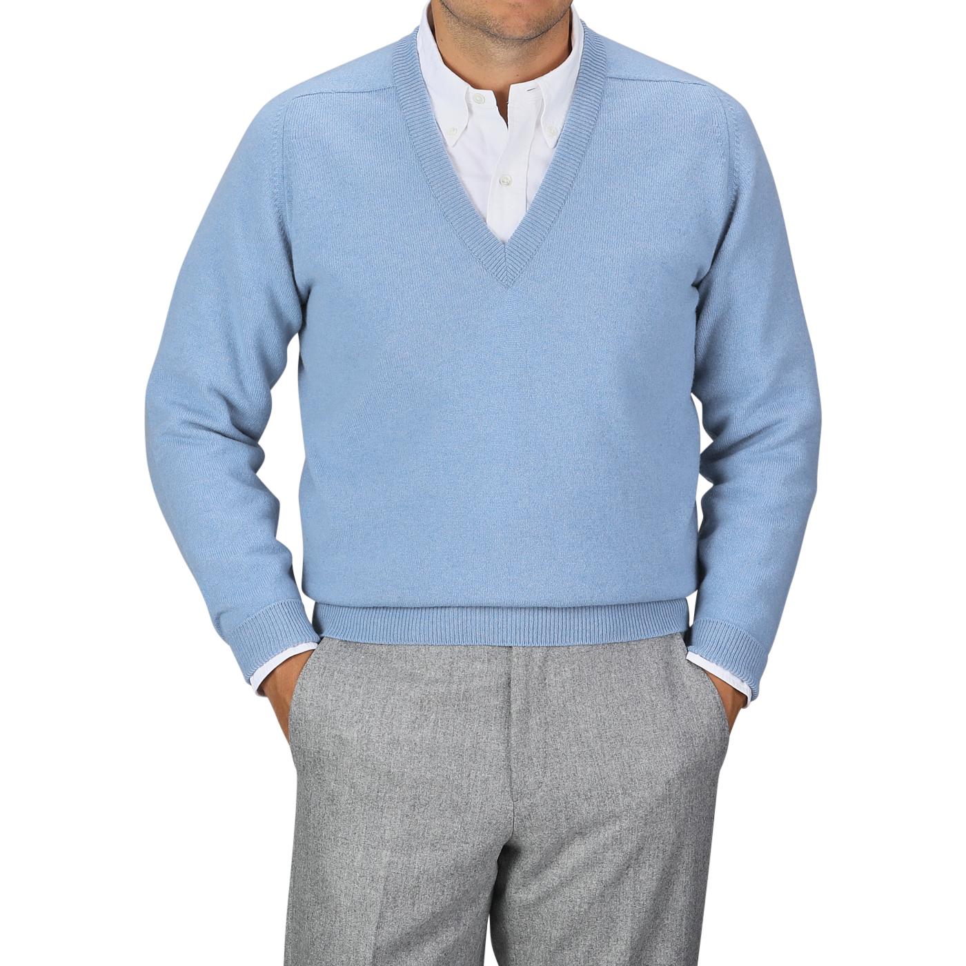 An individual donning an Iceberg Blue Deep V-Neck Lambswool Sweater from William Lockie over a white collared shirt and gray pants stands with their hands in their pants pockets against a plain gray background.