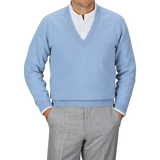 An individual donning an Iceberg Blue Deep V-Neck Lambswool Sweater from William Lockie over a white collared shirt and gray pants stands with their hands in their pants pockets against a plain gray background.