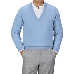 An individual donning an Iceberg Blue Deep V-Neck Lambswool Sweater from William Lockie over a white collared shirt and gray pants stands with their hands in their pants pockets against a plain gray background.