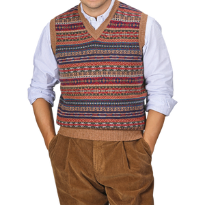 A person wearing a William Lockie Light Brown Fair Isle V-Neck Lambswool Slipover over a blue and white striped shirt, with hands in the pockets of brown corduroy pants.