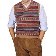 A person wearing a William Lockie Light Brown Fair Isle V-Neck Lambswool Slipover over a blue and white striped shirt, with hands in the pockets of brown corduroy pants.