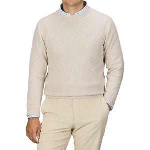 A person is wearing a William Lockie Linen Beige Crew Neck Lambswool Sweater over a light blue collared shirt, with matching beige pants, standing against a plain grey background.