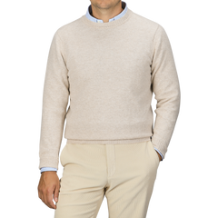 A person is wearing a William Lockie Linen Beige Crew Neck Lambswool Sweater over a light blue collared shirt, with matching beige pants, standing against a plain grey background.