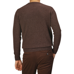 A person seen from the back wearing a mocha brown Mocha Brown Crew Neck Lambswool Sweater by William Lockie and brown pants, standing against a plain gray background. The knitwear fashion highlights the rich texture of the Scottish lambswool fabric.