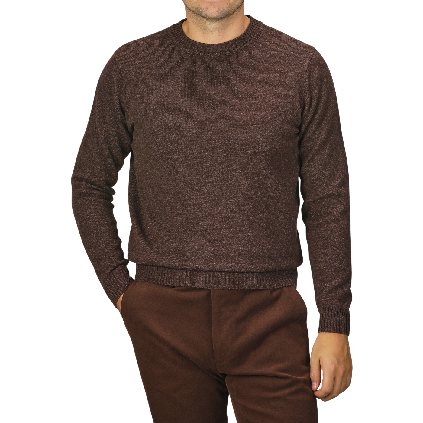 A person wearing a William Lockie Mocha Brown Crew Neck Lambswool Sweater and matching brown pants is standing with hands partially in pockets. The background is plain and gray.