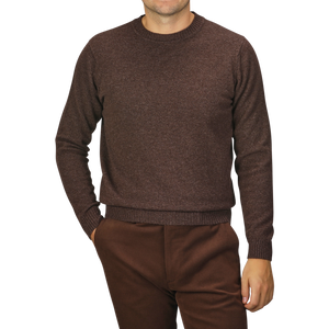 A person wearing a William Lockie Mocha Brown Crew Neck Lambswool Sweater and matching brown pants is standing with hands partially in pockets. The background is plain and gray.