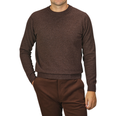 A person wearing a William Lockie Mocha Brown Crew Neck Lambswool Sweater and matching brown pants is standing with hands partially in pockets. The background is plain and gray.