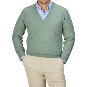 A person in a Mojito Green Deep V-Neck Lambswool Sweater by William Lockie over a blue collared shirt with beige pants, standing with hands in pockets against a gray background.