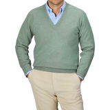 A person in a Mojito Green Deep V-Neck Lambswool Sweater by William Lockie over a blue collared shirt with beige pants, standing with hands in pockets against a gray background.