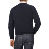 A man wearing a Navy Blue Deep V-Neck Lambswool Sweater by William Lockie and denim jeans is shown from the back against a plain gray background.