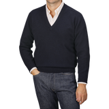 A person dressed in a navy blue deep V-neck lambswool sweater by William Lockie over a collared shirt and jeans, standing with one hand in their pocket against a neutral background.