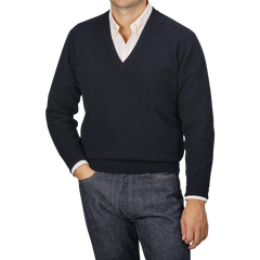 A person dressed in a navy blue deep V-neck lambswool sweater by William Lockie over a collared shirt and jeans, standing with one hand in their pocket against a neutral background.