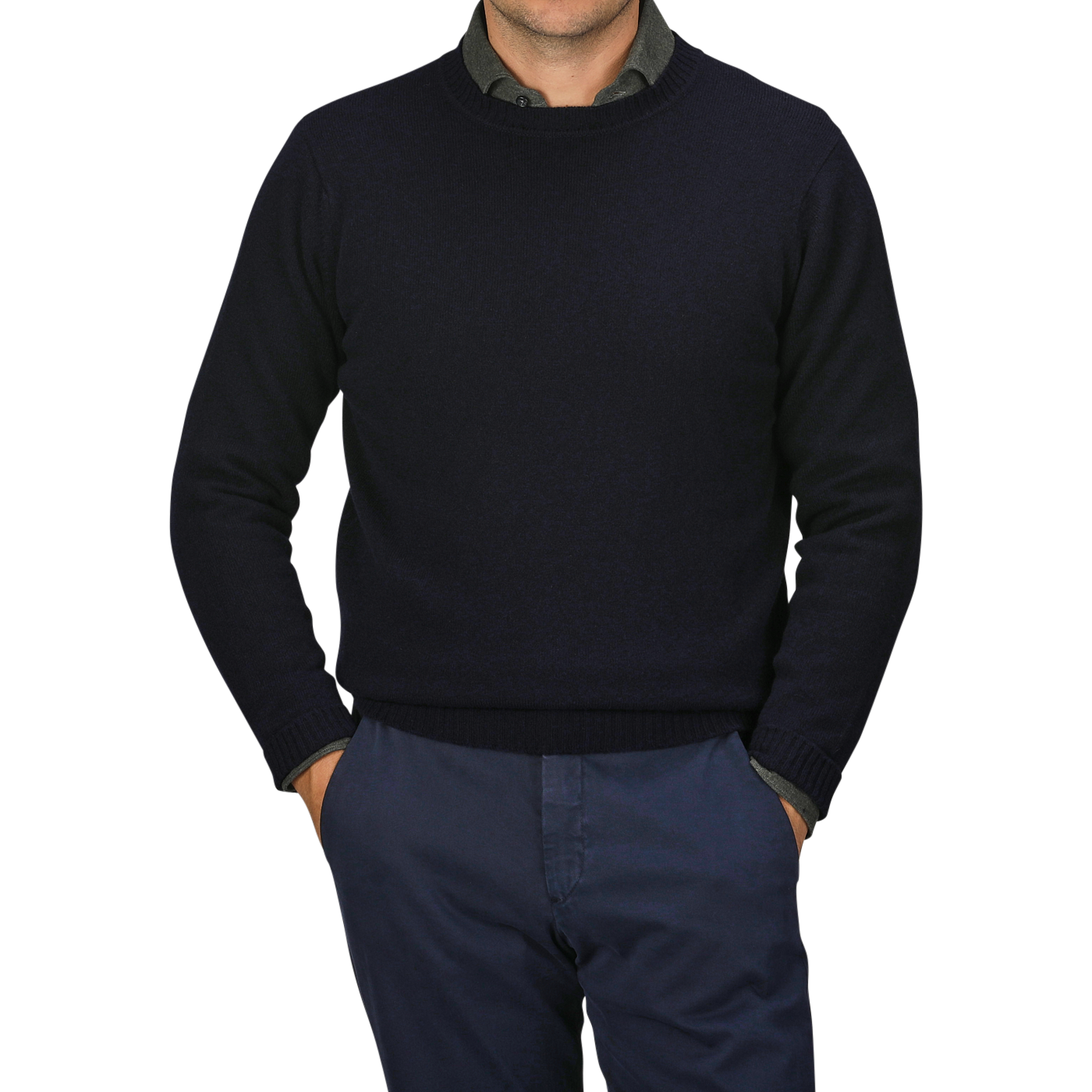 A man wearing a dark William Lockie Navy Crewneck Lambswool Sweater and dark blue pants stands with his hands in his pockets against a grey background, epitomizing smart casual elegance.