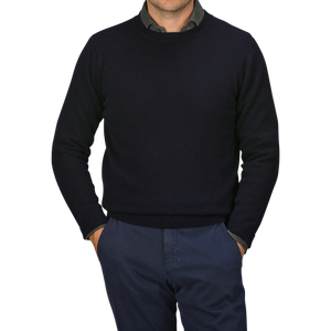 A man wearing a dark William Lockie Navy Crewneck Lambswool Sweater and dark blue pants stands with his hands in his pockets against a grey background, epitomizing smart casual elegance.