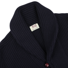 Close-up of the William Lockie Navy Lambswool Shawl Collar Cardigan, showcasing its dark blue ribbed design and luxurious Scottish lambswool fabric. The cardigan features distinctive leather buttons, with one red button visible near the bottom.