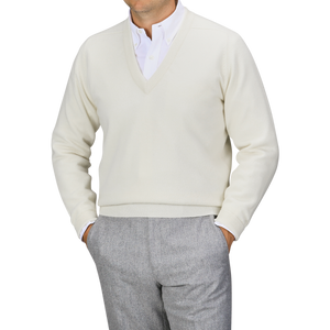 A person is wearing an off-white William Lockie Off-White Deep V-Neck Lambswool Sweater over a white collared shirt and gray trousers. The pose is casual with hands in their pockets.