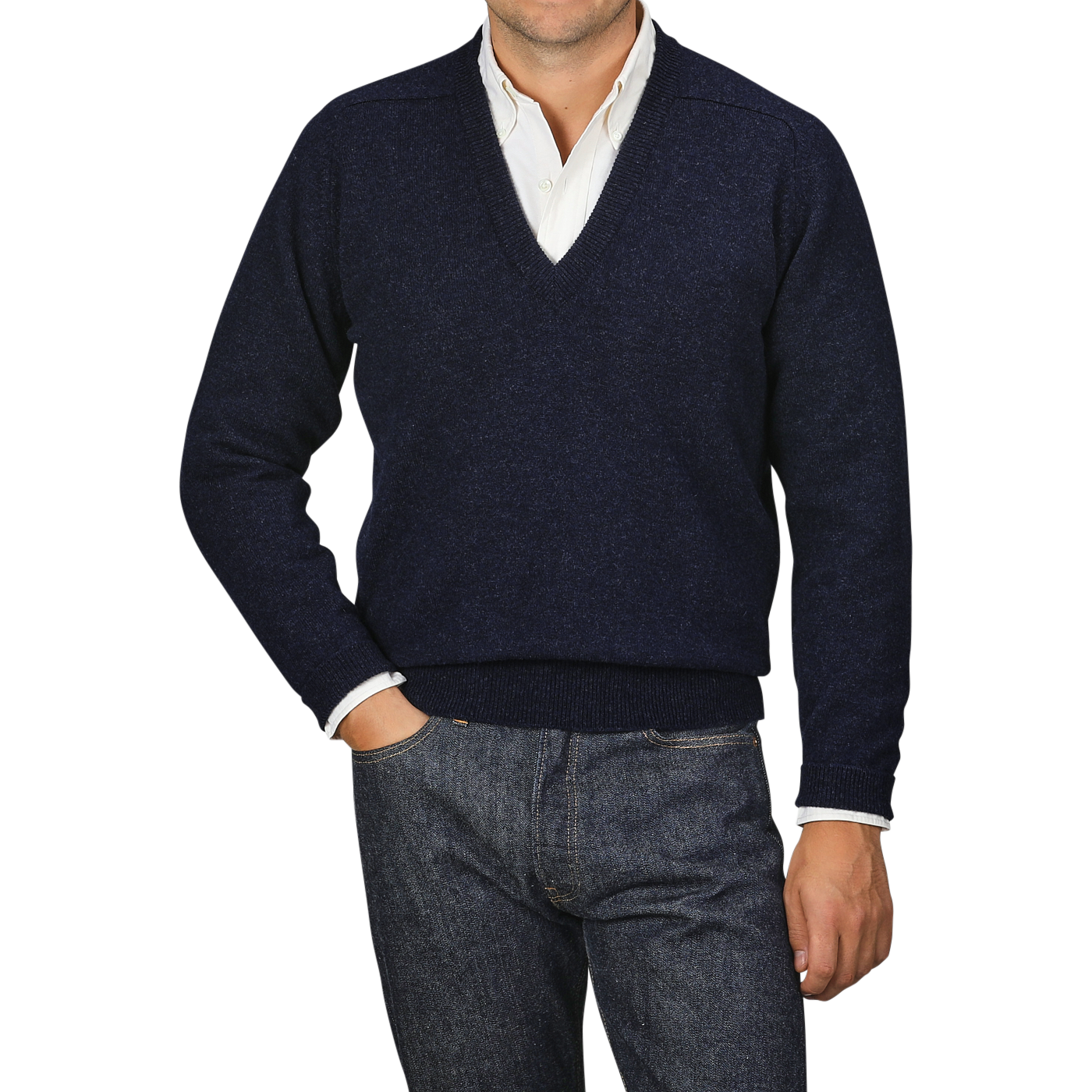 A person wearing an Oxford Blue Deep V-Neck Lambswool Sweater by William Lockie over a white collared shirt, with hands in the pockets of dark blue jeans, stands against a plain gray background.