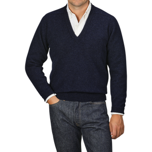 A person wearing an Oxford Blue Deep V-Neck Lambswool Sweater by William Lockie over a white collared shirt, with hands in the pockets of dark blue jeans, stands against a plain gray background.