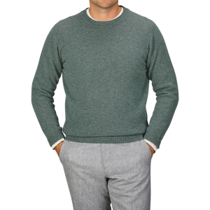 A person wearing a Peacock Crew Neck Lambswool Sweater by William Lockie and grey pants stands with hands in pockets against a plain grey background.