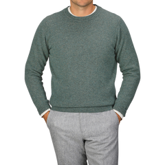 A person wearing a Peacock Crew Neck Lambswool Sweater by William Lockie and grey pants stands with hands in pockets against a plain grey background.