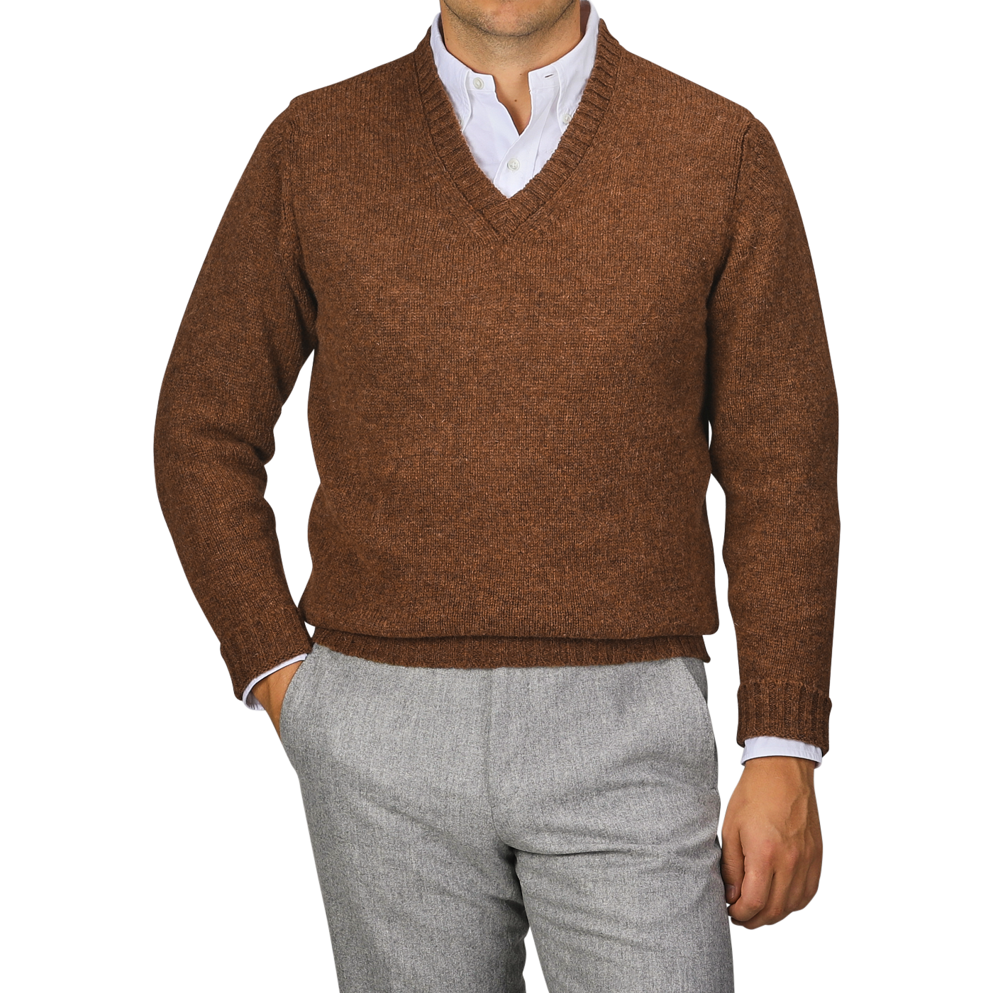 A person wearing a Pecan Brown Shetland Lambswool V-Neck Sweater from William Lockie over a white collared shirt and grey pants stands with one hand in their pocket against a plain background.