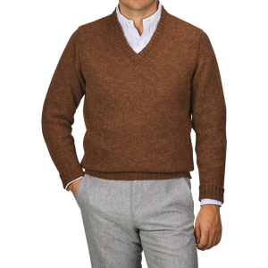 A person wearing a Pecan Brown Shetland Lambswool V-Neck Sweater from William Lockie over a white collared shirt and grey pants stands with one hand in their pocket against a plain background.