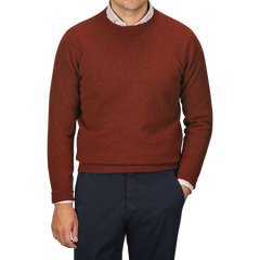 A person wearing a William Lockie Sienna Brown Crew Neck Lambswool Sweater over a white collared shirt pairs them with dark trousers, standing in front of a plain, light grey background.