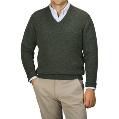 A man stands with hands in pockets, donning a William Lockie Spruce Brushed Wool Shetland Lambswool V-Neck sweater over a light blue shirt, paired with khaki pants.