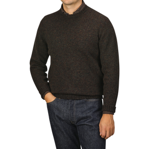 A person wearing the Turin Brushed Shetland Lambswool Crew Neck sweater from William Lockie in dark brown and blue jeans stands against a gray background, casually placing their left hand in their pocket.