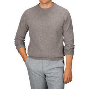 A person wearing a William Lockie Vole Beige Crew Neck Lambswool Sweater and gray pants, standing with one hand in a pocket against a plain background.