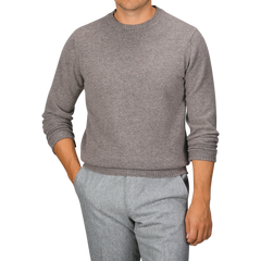 A person wearing a William Lockie Vole Beige Crew Neck Lambswool Sweater and gray pants, standing with one hand in a pocket against a plain background.