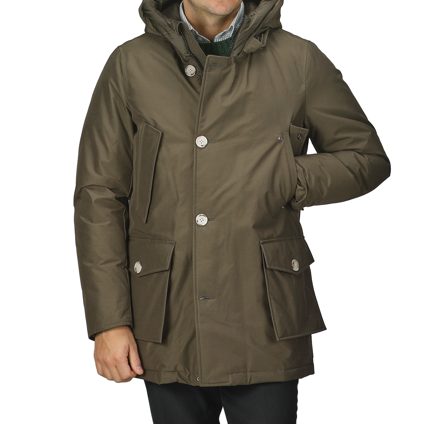 A person wearing a dark green Arctic Parka by Woolrich, featuring a hood, stretch nylon fabric, buttons, and multiple pockets, standing with one hand tucked in a pocket.