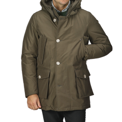 A person wearing a dark green Arctic Parka by Woolrich, featuring a hood, stretch nylon fabric, buttons, and multiple pockets, standing with one hand tucked in a pocket.