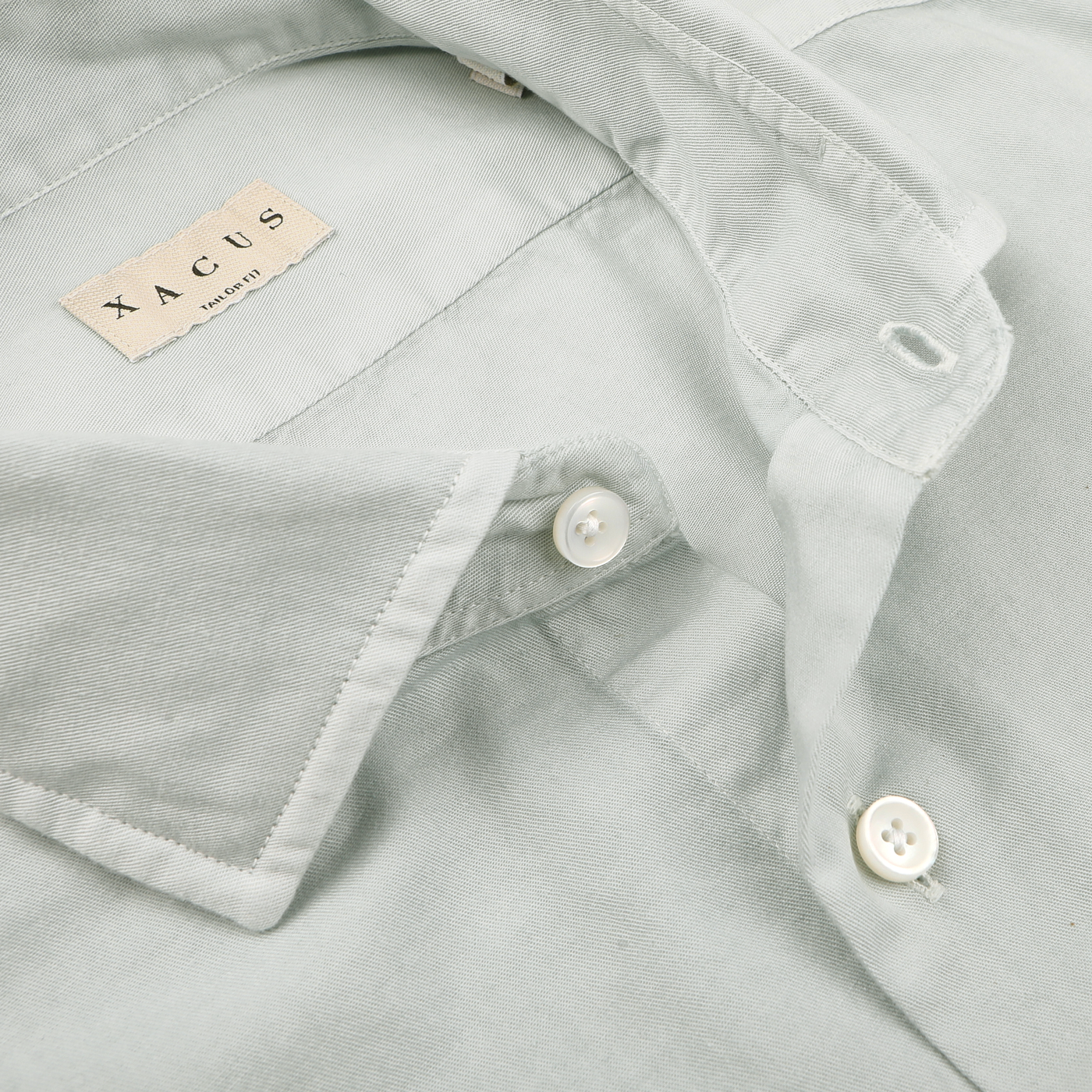 Close-up of a sage green shirt with white buttons, a cut-away collar, and a visible label reading "Xacus.