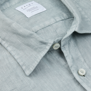 Close-up of a Xacus Sage Green Washed Linen Legacy Shirt with visible texture, a label, and buttons.