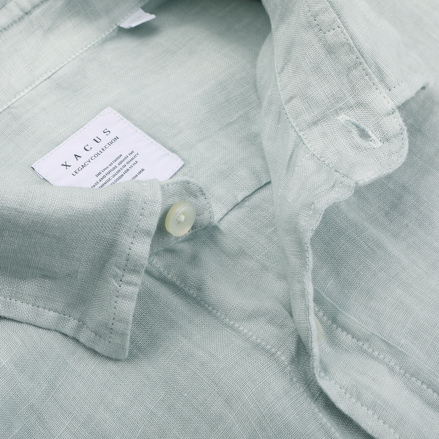 Close-up of a sage green washed linen Legacy shirt by Xacus, featuring a buttoned cut-away collar. A label inside reads "Xacus Linen Collection.