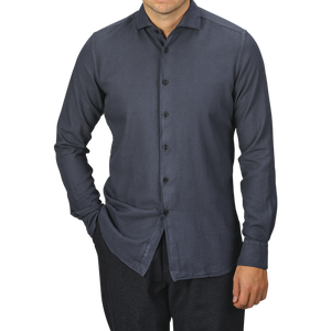 A man is wearing a Washed Navy Mussola Cotton Tailor Fit Shirt from Xacus. The pure cotton material and slim fit accentuate his frame as he stands with one hand in his pocket against a plain background. His face is not visible.