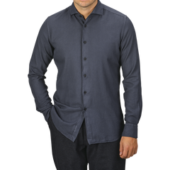 A man is wearing a Washed Navy Mussola Cotton Tailor Fit Shirt from Xacus. The pure cotton material and slim fit accentuate his frame as he stands with one hand in his pocket against a plain background. His face is not visible.